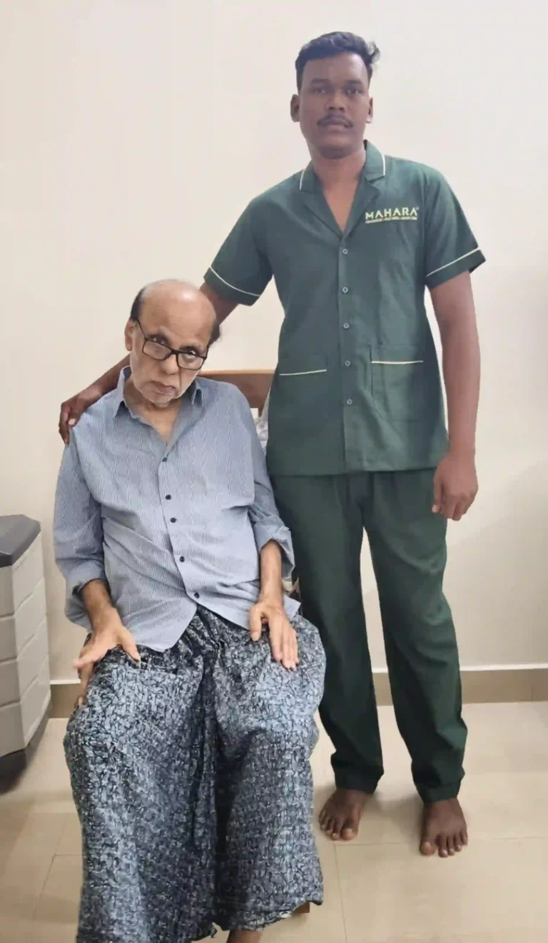 Male Mahara home nurse standing beside an elderly male patient seated on a chair